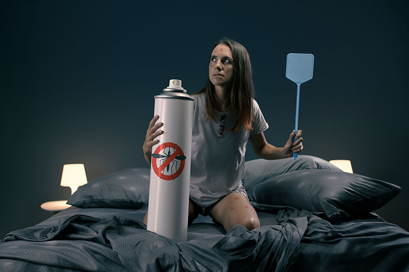A woman is sitting on a bed in her bedroom with a huge mosquito repellent spray in one hand and a fly swatter in another hand.