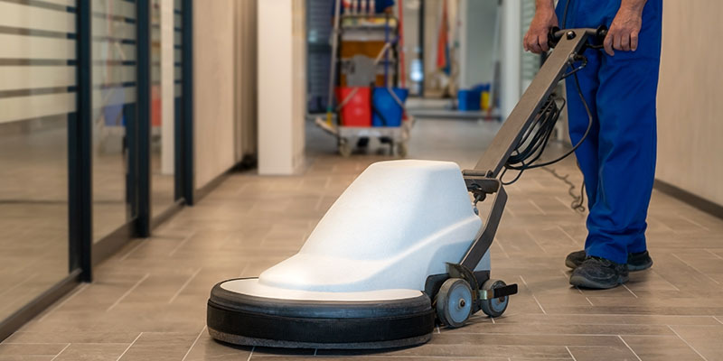 A house cleaning professional is polishing the hard floor with the help of movable cleaning machine in an office.
